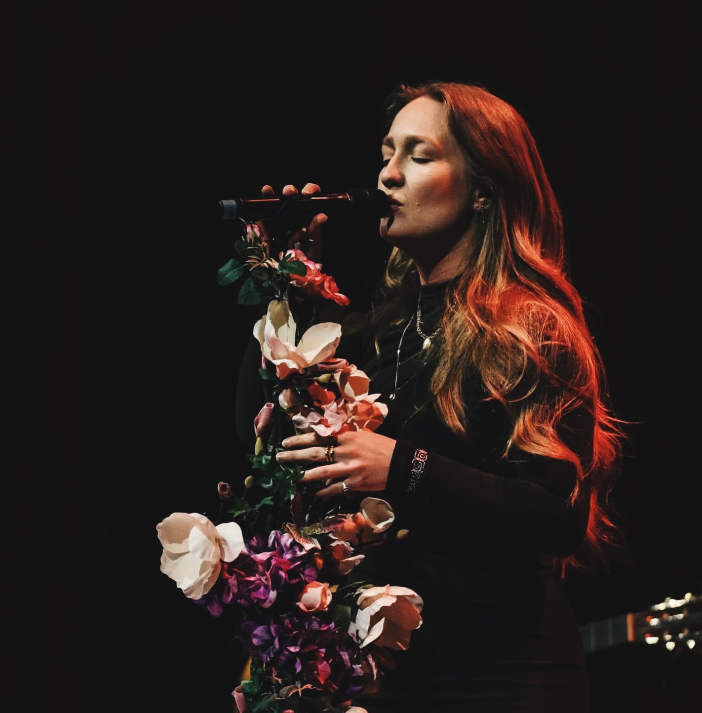 Vilma Olsson singing on a dark stage, holding flowers in warm red light.